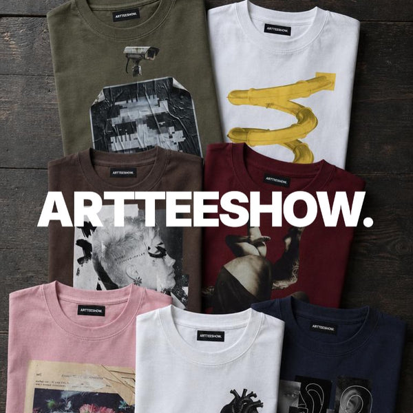 Seven folded ARTTEESHOW. graphic t-shirts in various colors (olive green, white, brown, burgundy, pink, white, and navy) are arranged in a grid on a dark wood background, with a large white 'ARTTEESHOW.