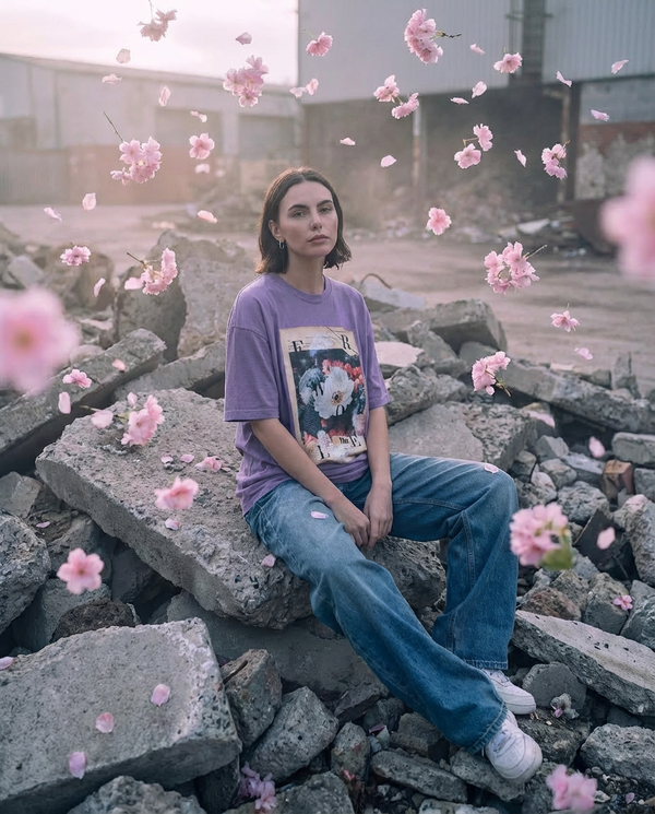 Candid High-fashion editorial streetwear photography. Full body shot. The female model is sitting on a pile of grey concrete rubble, but she is surrounded by floating pink flowers. She wears the Washed Purple Oversized 260gsm T-Shirt OSAKA SIX: 004 FLORA MORTIS