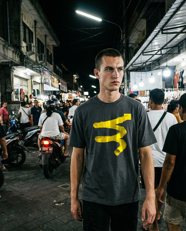 Candid editorial cinematic streetwear photography. The male model is walking through a busy street in Bali. He is wearing the Dark Grey 260gsm Heavyweight T-Shirt with the bold yellow conduit graphic. The yellow graphic reflects the overhead fluorescent overhead street lights.