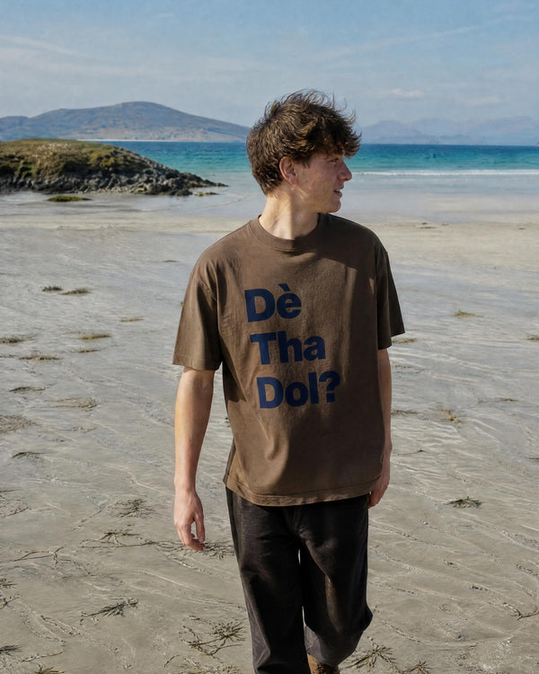A man wearing the ldark brown ARTTEESHOW Dè Tha Dol drop shoulder t-shirt in 260GSM heavyweight cotton, walking across wet sand on a wide Hebridean beach in the Outer Hebrides with the Scottish Gaelic phrase printed in navy across the chest.