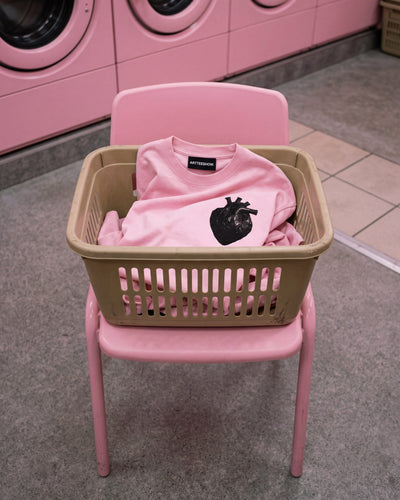 Azalea pink 260GSM heavyweight t-shirt 
naturally discarded in translucent pink 
plastic laundry basket on pink chair. 
Anatomical bin bag heart graphic facing 
upward. ARTTEESHOW. woven neck label visible. 
Pink plastic toys alongside. Laundrette setting. 
ARTTEESHOW. Heavy Duty: The Idol. 
Limited edition graphic tee UK.