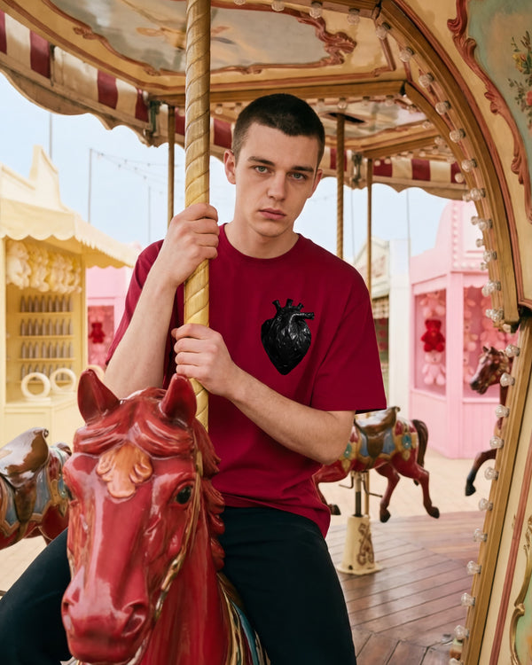 ARTTEESHOW Heavy Duty The Novelty 260GSM cardinal red boxy fit graphic tee worn by male model at day time European fairground carousel, bin bag heart graphic visible on left chest, Heavy Duty Collection limited to 100 pieces.