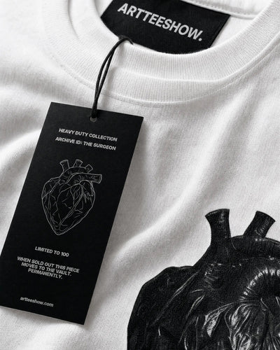 Extreme macro detail of the heavyweight 260gsm cotton tee The Surgeon, the inner neck label and the branded hang tag in sharp focus resting on the dense combed cotton weave, the edge of the matte black heart print soft behind. Soft even lighting, shallow depth, premium tactile product photography, true to fabric.