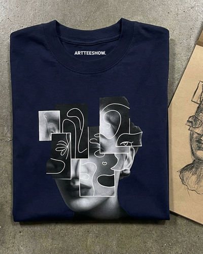 Isolde navy tee folded on concrete beside a sketchbook, showing the black and white cubist face collage graphic.