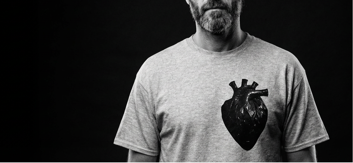 Cinematic black and white hero banner for the Heavy Duty streetwear collection by ArtTeeShow. Features a stoic portrait of the founder wearing sunglasses and the Sport Grey heavyweight t-shirt with a realistic black bin bag anatomical heart graphic. High-fashion gritty aesthetic.