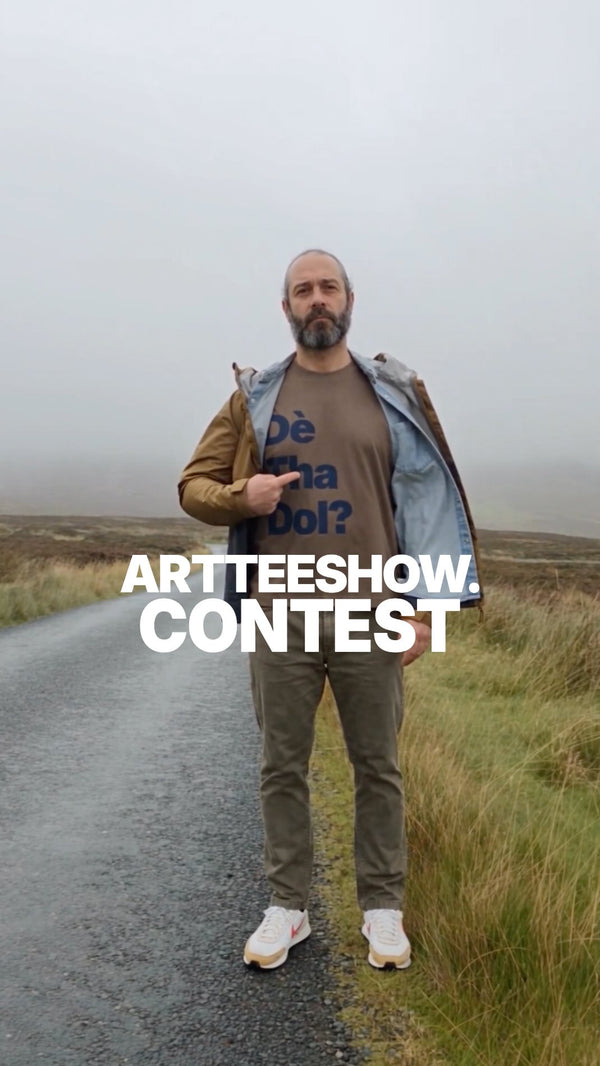 Laurent stands on a wet single-track road in the misty Outer Hebrides, wearing an open outdoor jacket over a dark brown “Dè Tha Dol?” T-shirt and pointing to the design, with rugged hills, moorland and a loch visible in the background.