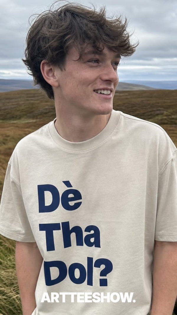 A young man in scottish moorlands wearing a cream-colored t-shirt with the text 'Dè Tha Dol?' in dark blue and 'ARTTEESHOW.' at the bottom.