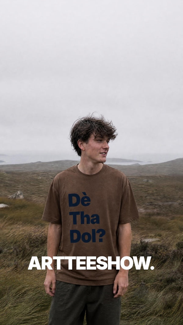 A man standing outdoors in a grassy, hazy landscape wearing a brown graphic t-shirt with the text 'Dé Tha Dol?' in navy blue and 'ARTTEESHOW.' in white at the bottom.