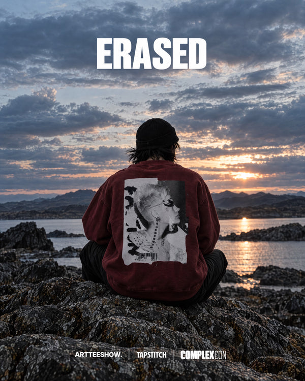 A person sits on rocks by the ocean at sunset, viewed from behind, wearing a burgundy hoodie with a large black-and-white portrait graphic on the back. The word 'ERASED' appears in large white letters across the top, with 'ARTTEESHOW | TAPSTITCH | COMPLEX' at the bottom.