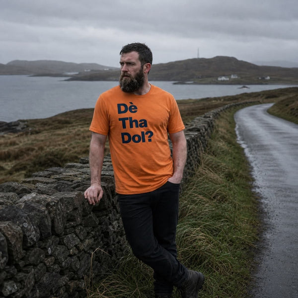 A bearded man wearing an orange t-shirt with the text 'Dé Tha Dol?' in navy blue, standing on a stone wall next to a wet road with a scottish coastal landscape in the background.